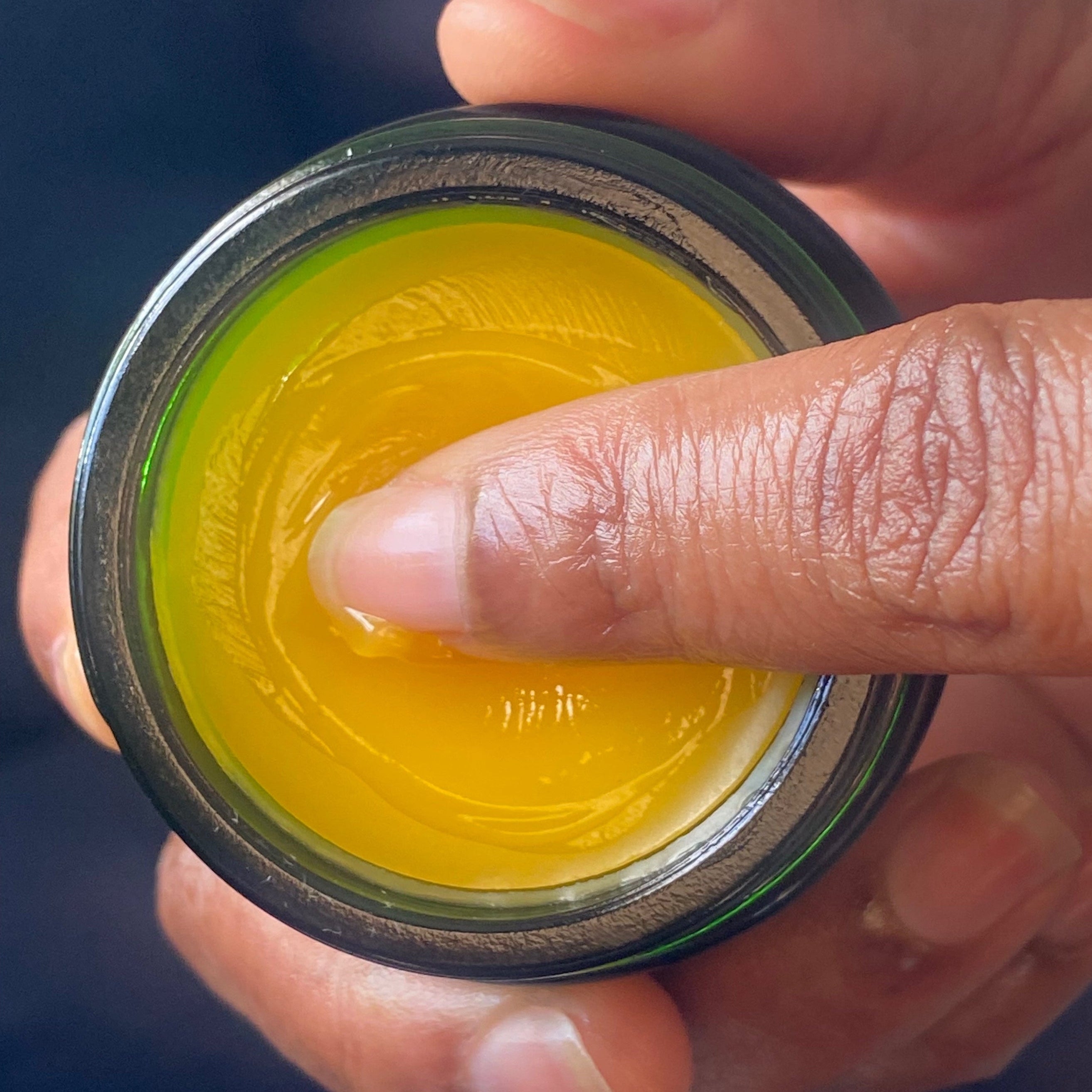 A person's finger dipping into an open jar of yellow ointment.
